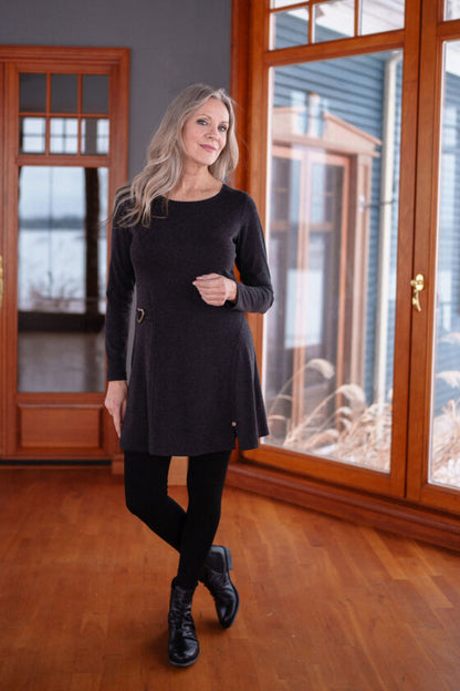 A woman wearing the Modigliani Tunic by Rien ne se Perd in Charcoal standing near a floor-to-ceiling window.