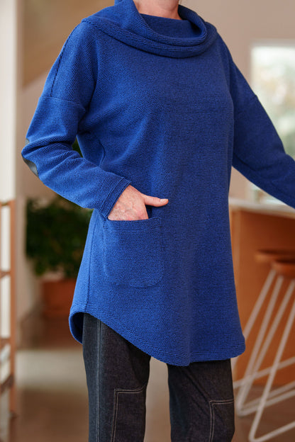 Close up view of a woman wearing the Myrique Tunic by Rien ne se Perd in Blue, an oversized chenille tunic with a large cowl neck, rounded hem, faux leather elbow patches, button placket at the left shoulder, and patch pocket on the front. She is wearing it with leggings and standing in a kitchen. 