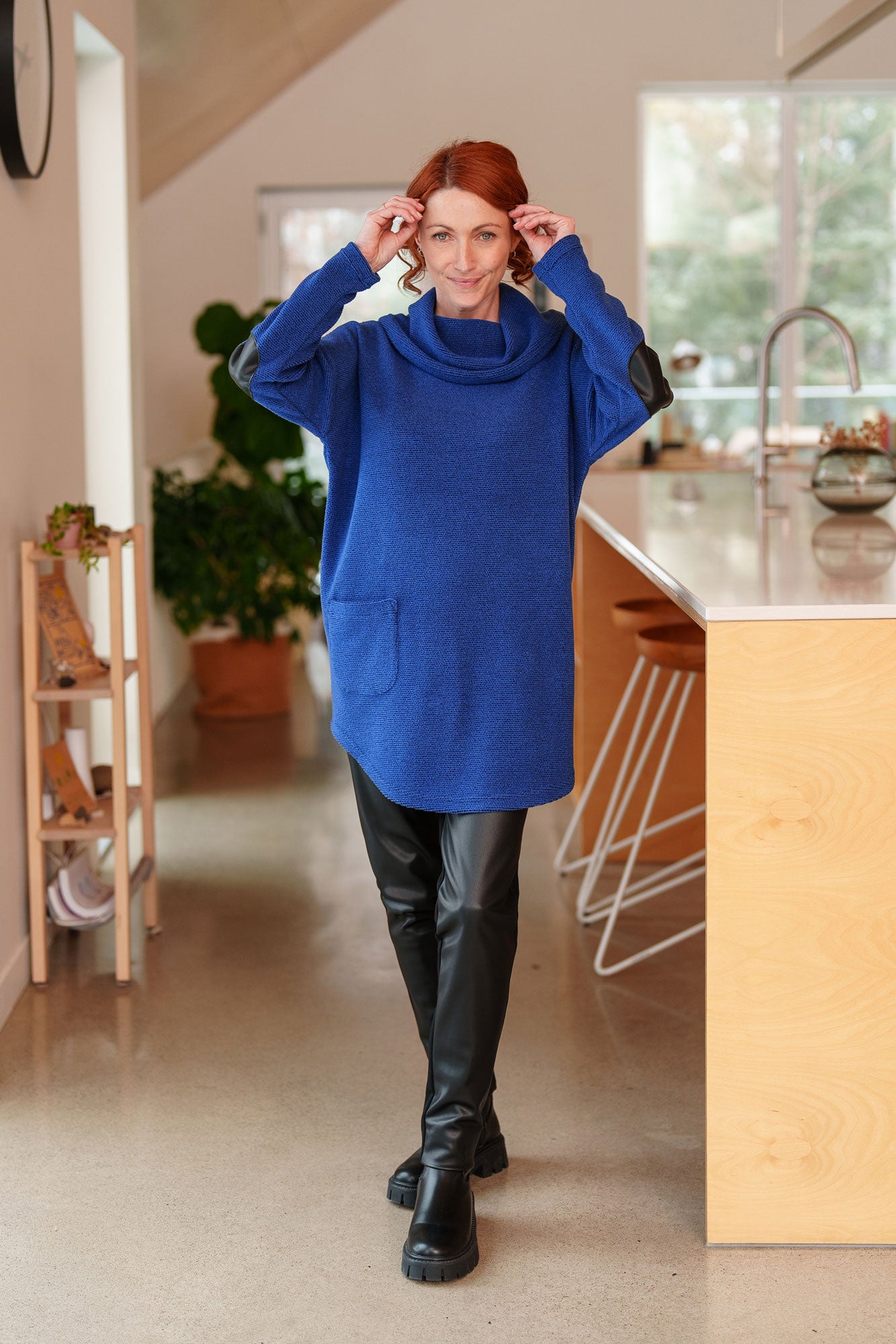 A woman wearing the Myrique Tunic by Rien ne se Perd in Blue, an oversized chenille tunic with a large cowl neck, rounded hem, faux leather elbow patches, button placket at the left shoulder, and patch pocket on the front. She is wearing it with the Resinosa Pants and standing in a kitchen. 