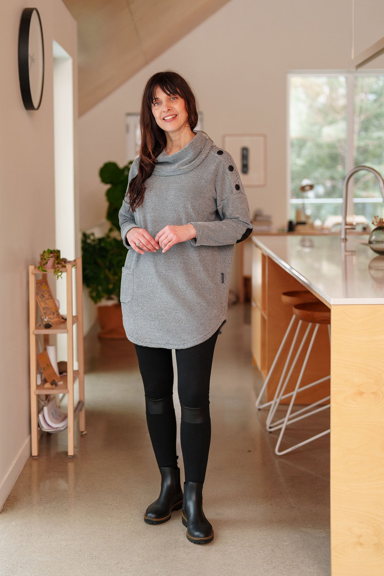 A woman wearing the Myrique Tunic by Rien ne se Perd in Grey, an oversized chenille tunic with a large cowl neck, rounded hem, faux leather elbow patches, button placket at the left shoulder, and patch pocket on the front. She is wearing it with leggings and standing in a kitchen. 