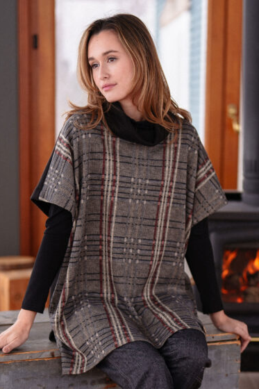 A woman wearing the Gauguin Sweater by Rien ne se Perd in Black, under the Borduas Tunic-Poncho in Checkered, sitting in front of a wood stove