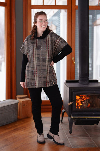 A woman wearing the Borduas Tunic-Poncho by Rien ne se Perd in a checkered print with black pants, standing beside a wood stove in a cabin.