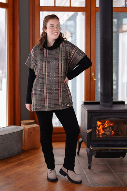 A woman wearing the Lichen Pant by Rien ne se Perd in Black, skinny leg pull-on pants in stretchy corduroy fabric. She is wearing them with plaid top and standing by a fireplace. 