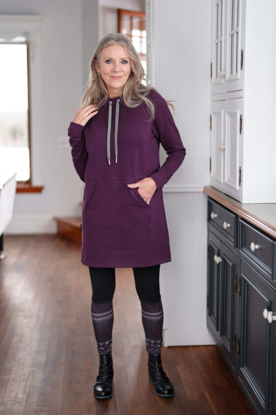 A woman wearing the Toulouse Tunic by Rien ne se Perd in Aubergine, standing beside a set of cupboards