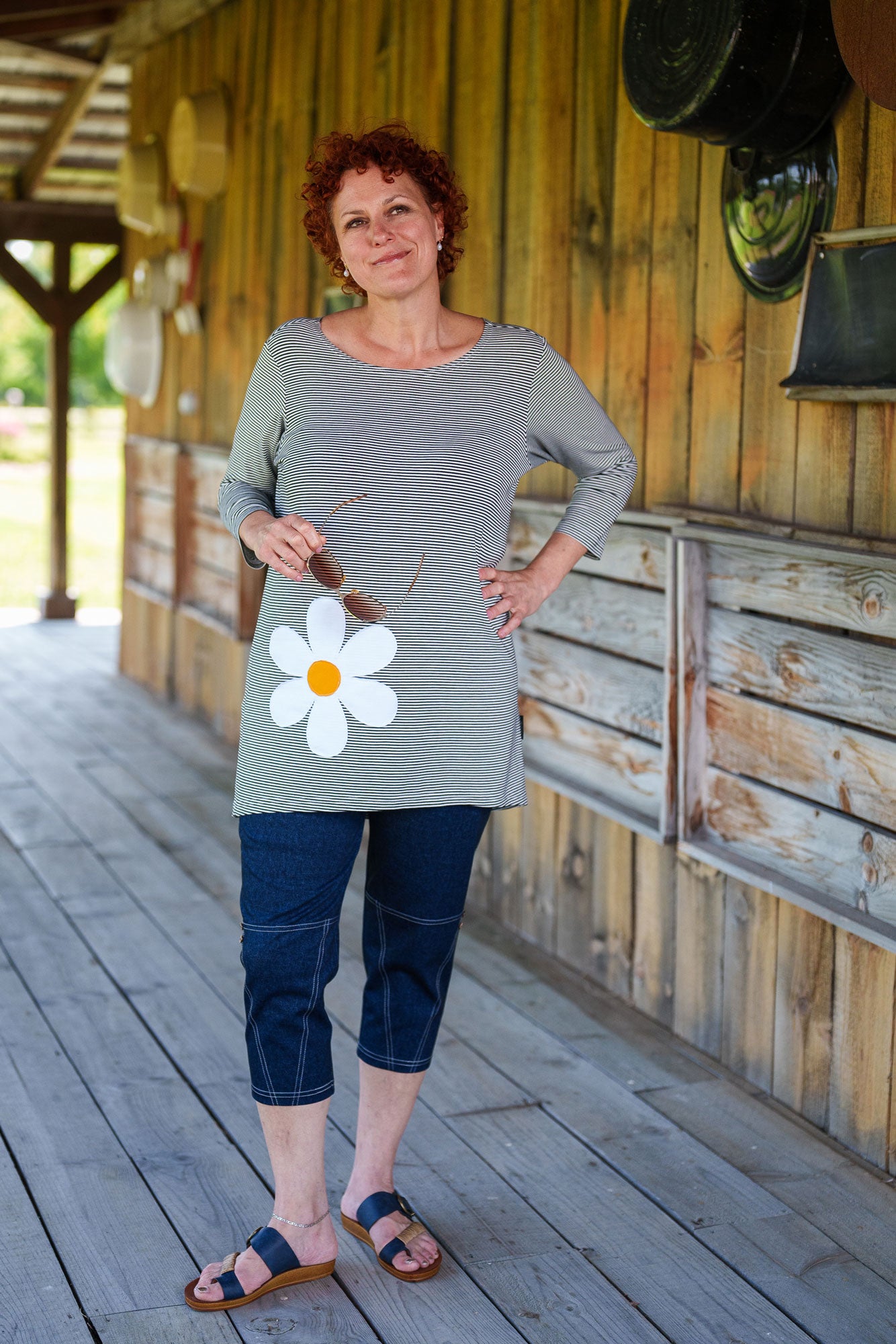 A woman wearing the Zumba Tunic by Rien ne se Perd, a striped navy and white piece with a boat neck, 3/4 sleeves, and a large daisy applique. She is wearing it with the LIndy Capri pants and standing outside a wooden building. 