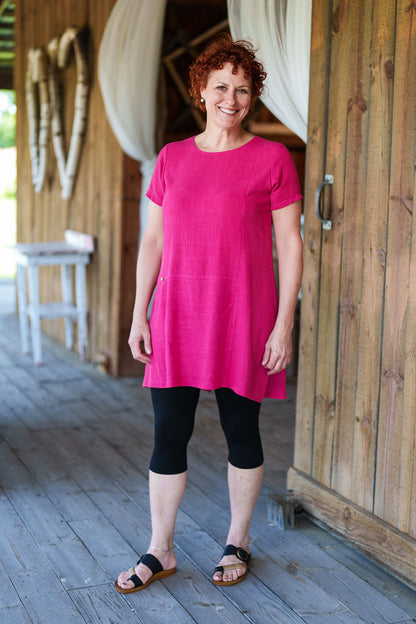 3/4 shot of a woman wearing the Merengue Tunic by Rien ne se Perd in Raspberry, a loose-fitting linen and viscose tunic with a round neck, short sleeves, asymmetrical cut, and patch pocket. She is wearing it with black capri leggings and is standing outside on a porch.