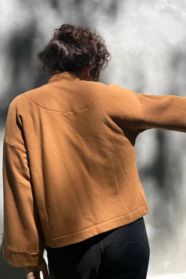 Back view of a woman wearing the Thuya Jacket by Kazak in Teak, standing in front of a grey wall 