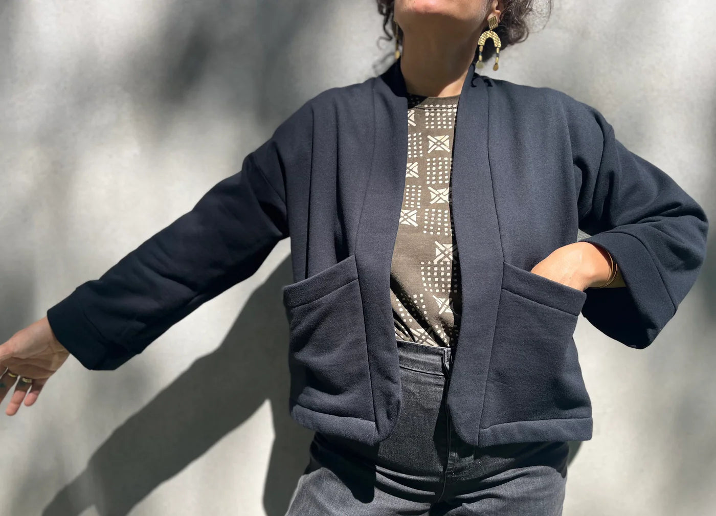 A woman wearing the Thuya Jacket by Kazak in Black, standing in front of a grey wall 