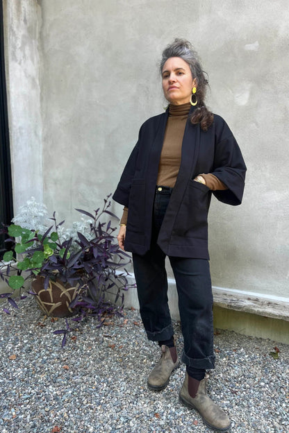 A woman wearing the Cydonia Cardigan by Kazak in Black, a hip-length open cardigan with 3/4 kimono sleeves and large patch pockets. She is wearing it with black pants and a black top and standing outside.