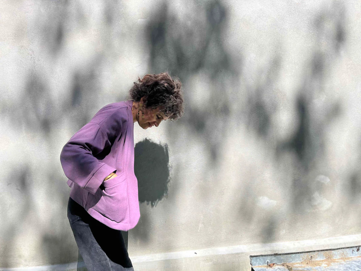 Side view of a woman wearing the Thuya Jacket by Kazak in Purple Haze, standing in front of a grey wall 