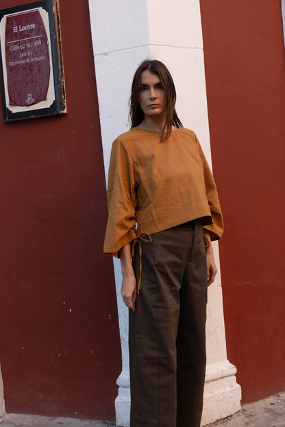 A woman wearing the Vienna Pants in Capers by MAS, high waisted pants with wide legs and a zipper and tab closure. She is wearing them with yellow top andstanding on the street. 