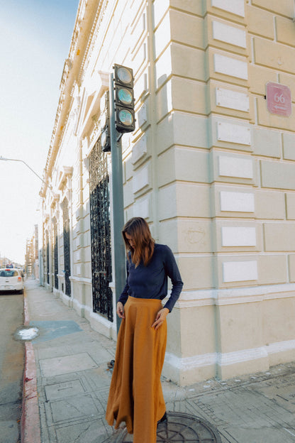 A woman wearing the Whistler Long Sleeve Top in Blueberry, a ribbed cotton knit with a round neck and straight cut. She is wearing it with a  brown skirt and standing against a light-colored building.