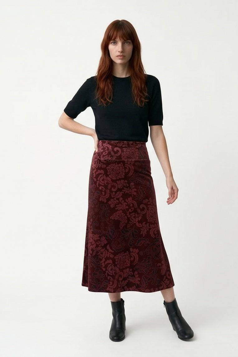 A woman wearing the Kollontai Winslet skirt, a burgundy velvet midi skirt, with a black top. She is standing against a plain white studio background.