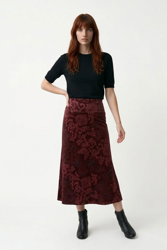 A woman wearing the Kollontai Winslet skirt, a burgundy velvet midi skirt, with a black top. She is standing against a plain white studio background.