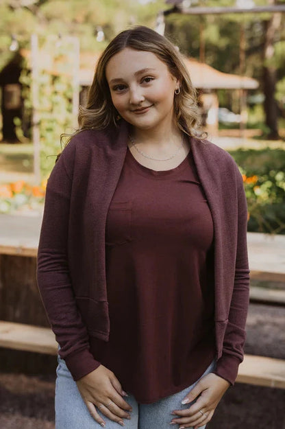 A woman wearing the Woodland Spruce Cardigan by Blondie in Berry, a short open front cardigan with long sleeves and a wide band across the bottom. She's wearing it with a t-shirt and jeans and standing outside. 