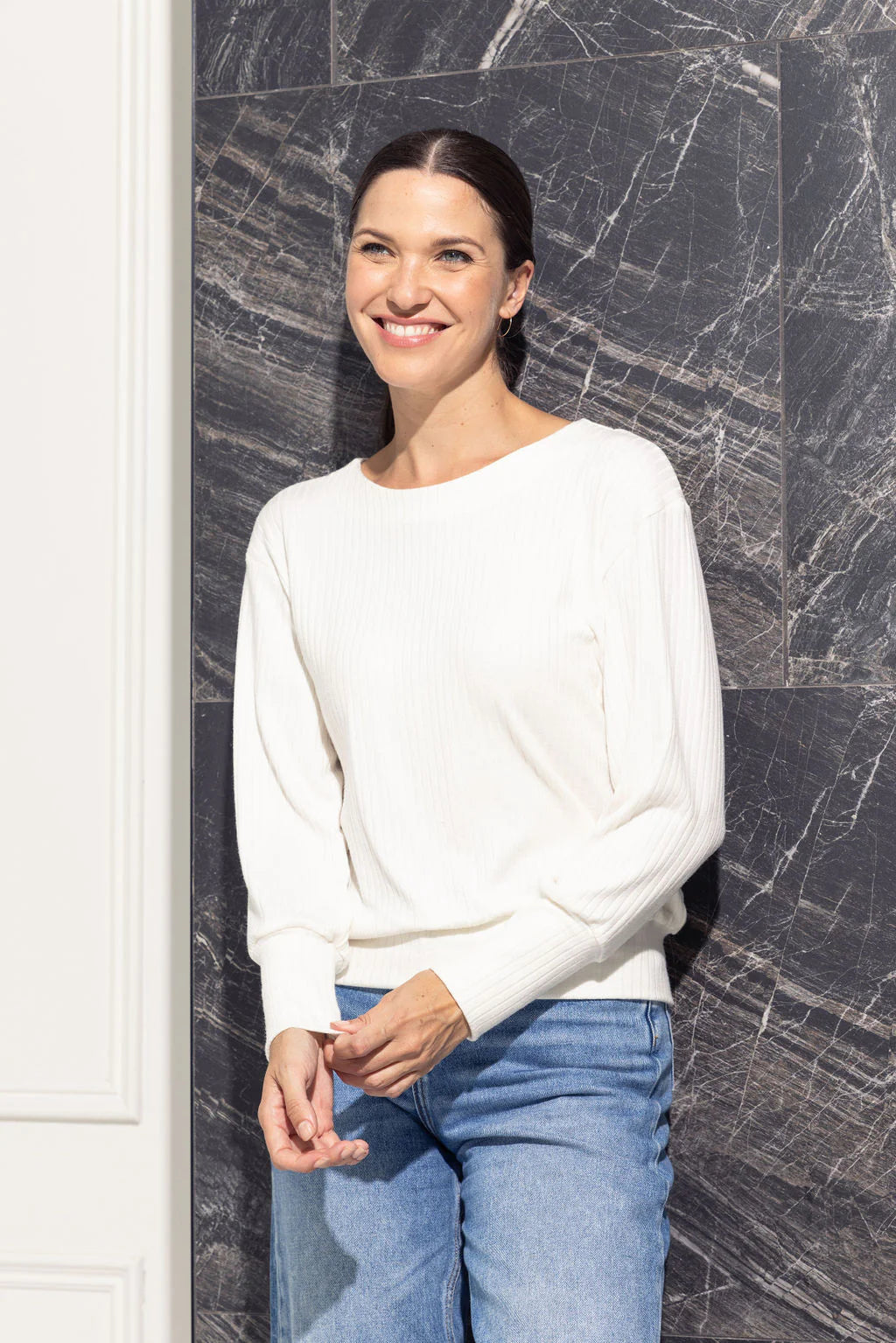 A woman wearing the Cherry Bobin Zeppelin Sweater in Cream with jeans, standing in front of a grey wall. The sweater has a round neck, a batwing shape, long cuffed sleeves and a fitted band across the bottom. 