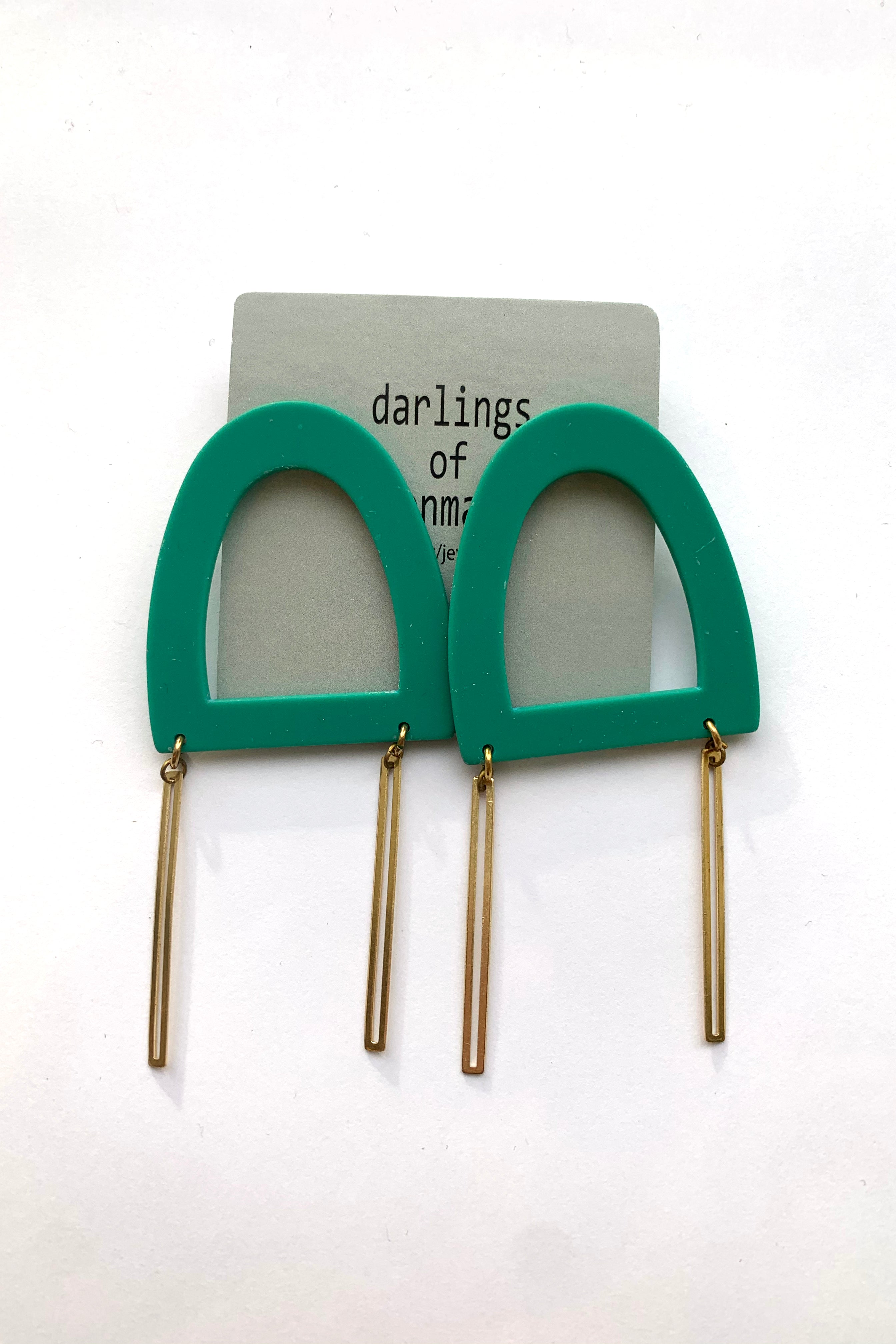 Lily earrings by Darlings of Denmark; large stud earrings with hollowed, acrylic, emerald geometric shape, and dangling raw brass sticks; flat lay
